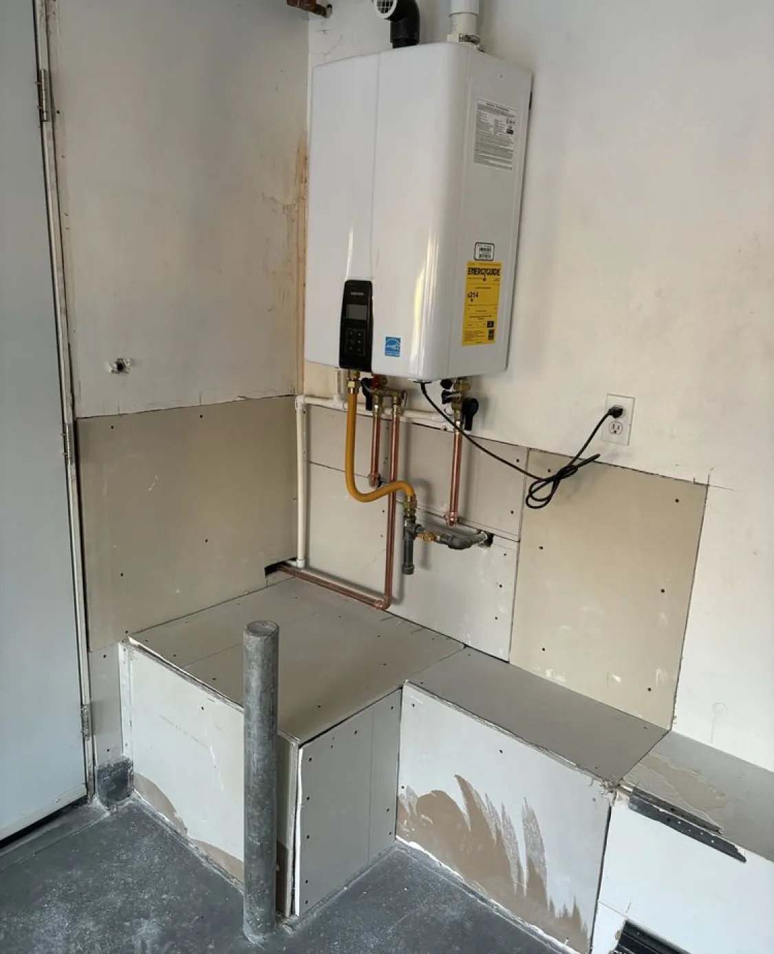 Tankless water heater mounted on a wall above unfinished drywall and plumbing in a utility area.
