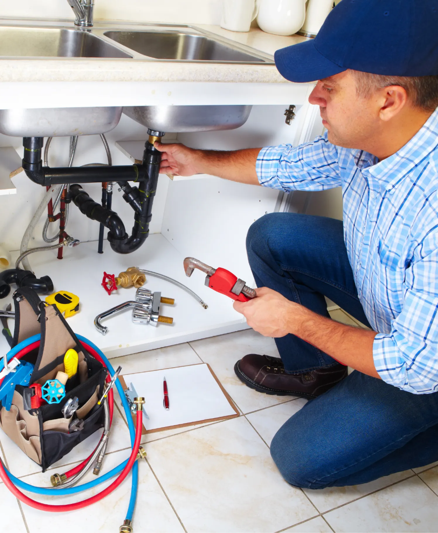 Plumber kneeling under a sink, repairing pipes with a wrench; toolbox and pipes on the floor nearby.