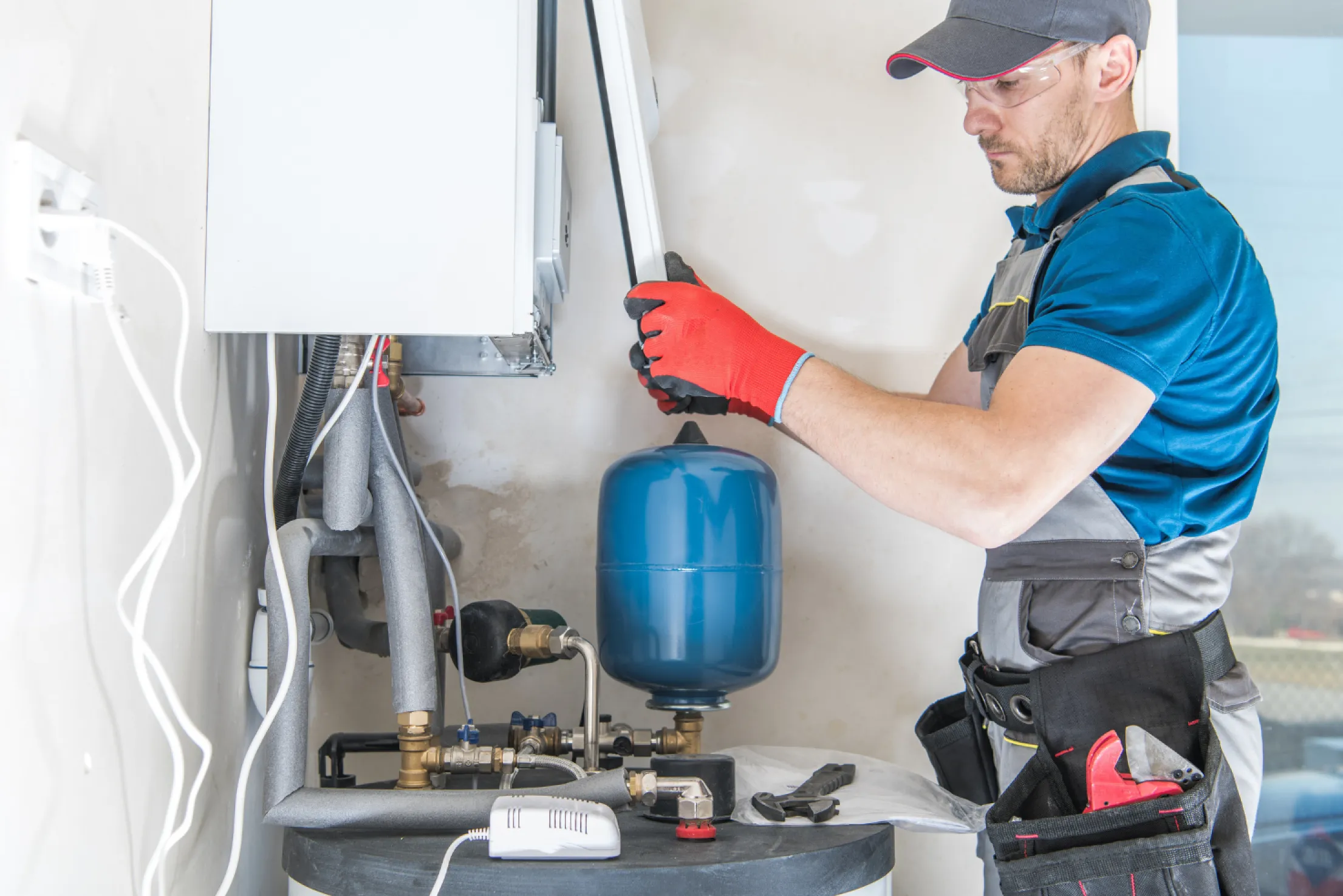 hero-1 Man in workwear and red gloves servicing a wall-mounted boiler with various pipes and a blue tank attached.