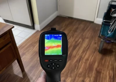 A hand holds a thermal imaging camera pointed at a wooden floor near a doorway.