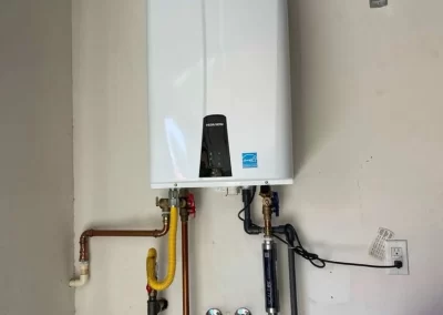 White tankless water heater mounted on a wall with various pipes and wires connected below it.