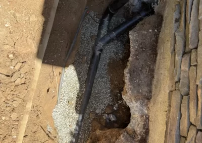 Black pipes run underground next to a stone wall with gravel and exposed soil around them.