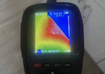 A hand holds a thermal imaging camera showing a heat map with temperature readings indoors.