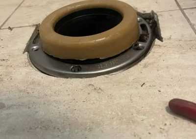 A wax toilet ring is installed on a metal flange on a tiled floor, with a red tool partially visible.