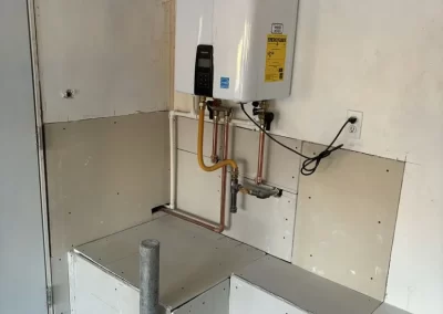 A tankless water heater mounted on a wall above unfinished drywall and exposed pipes in a utility area.