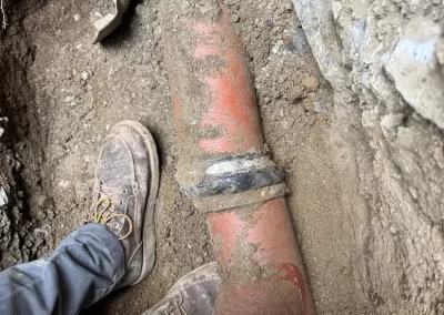 Two boots stand next to a large exposed clay pipe underground, with a shovel resting nearby in soil.