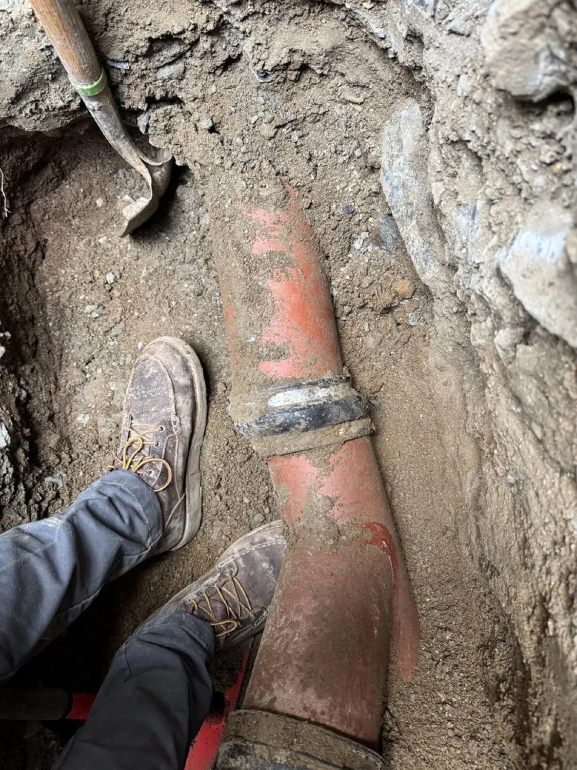 A person standing by an exposed underground pipe, surrounded by dirt and a shovel.