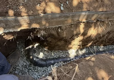 A black pipe runs underground in a trench filled with gravel, with a person's boot visible at the edge.