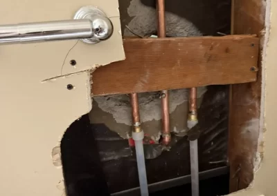 A large hole in a beige wall reveals copper pipes and wooden support behind drywall near a towel bar.