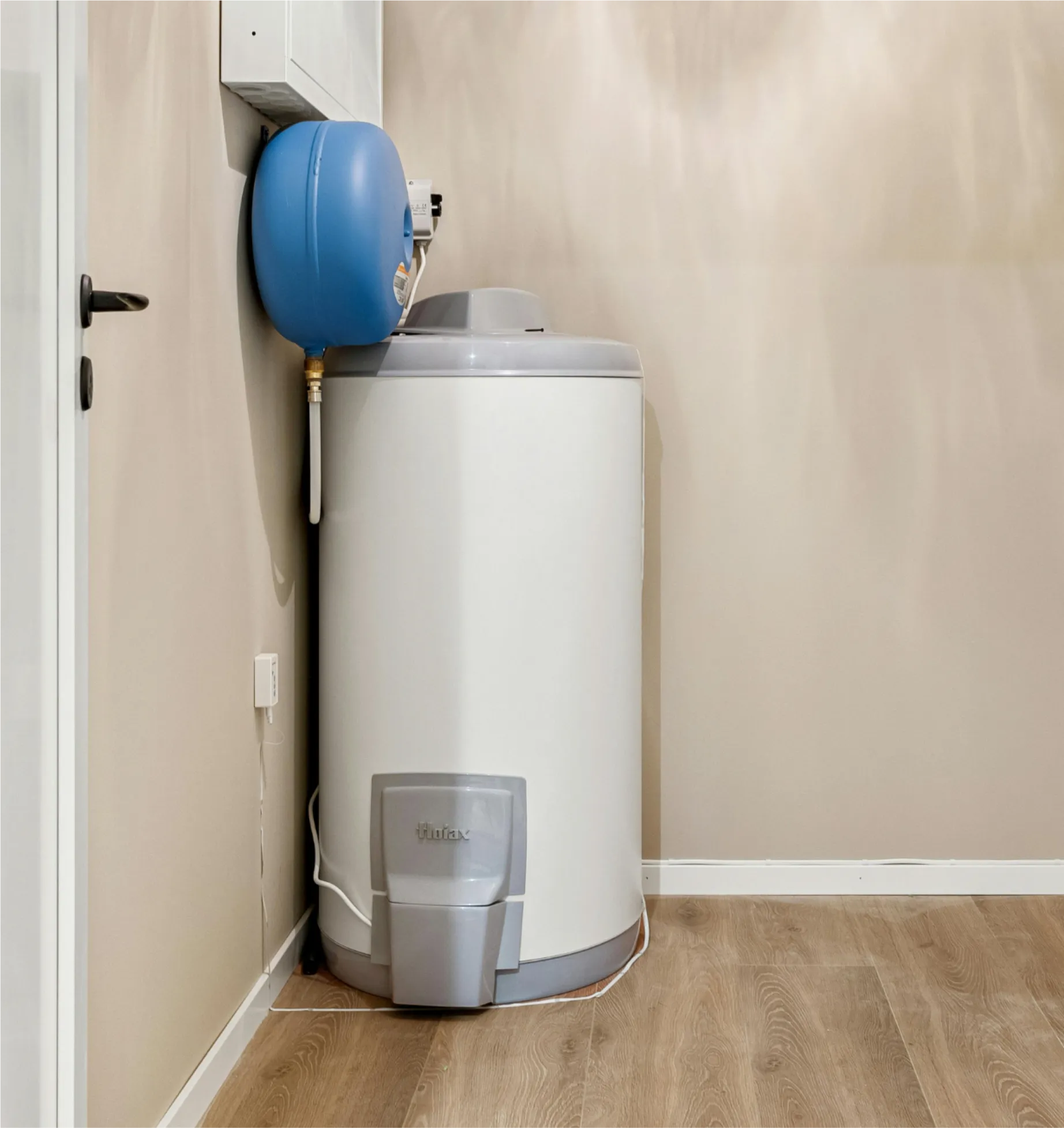 A white water heater with a blue expansion tank installed in a corner of a room with beige walls and wood flooring.