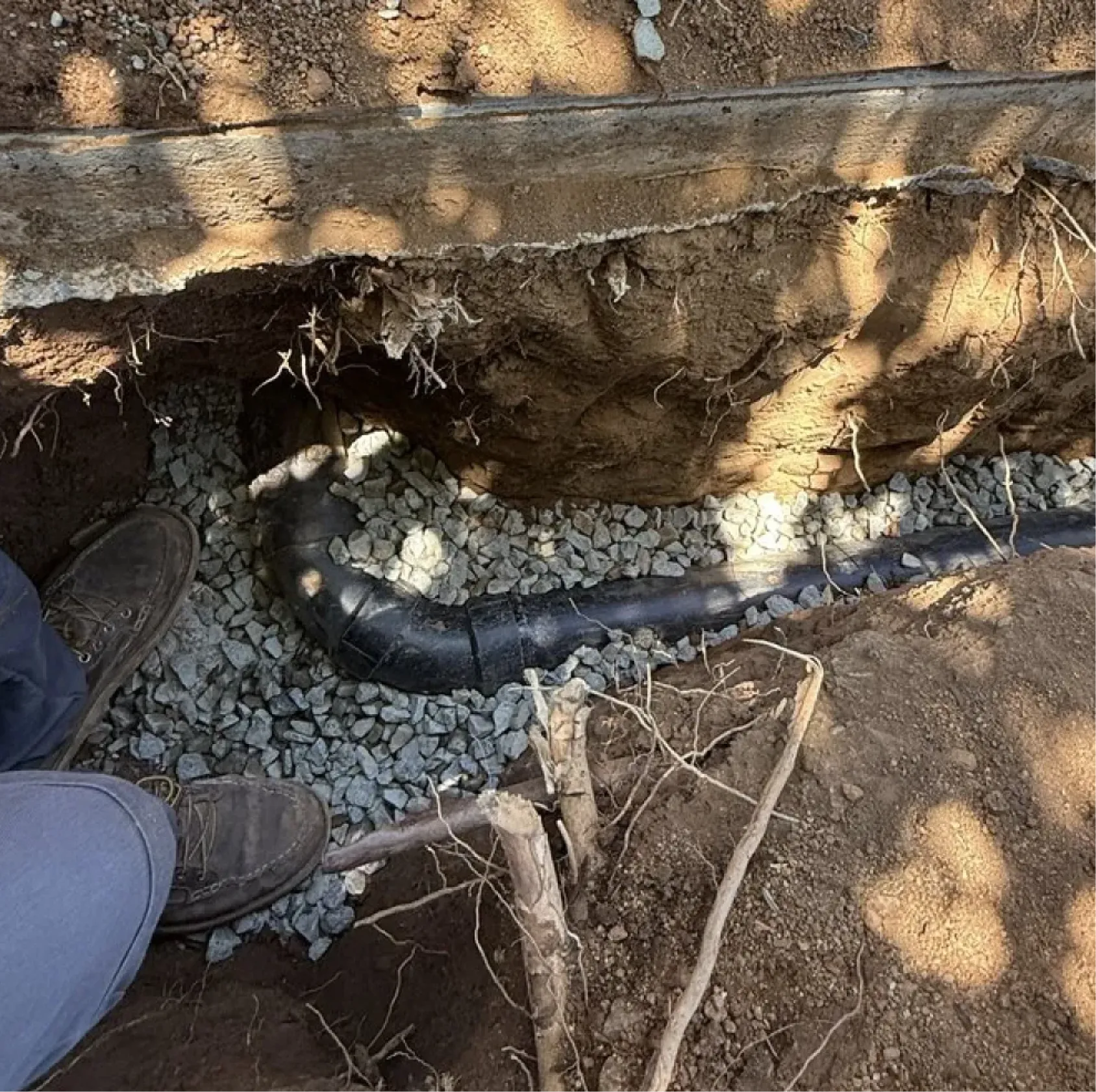 A person stands by a buried black pipe with a bend, surrounded by gravel in a dug trench.