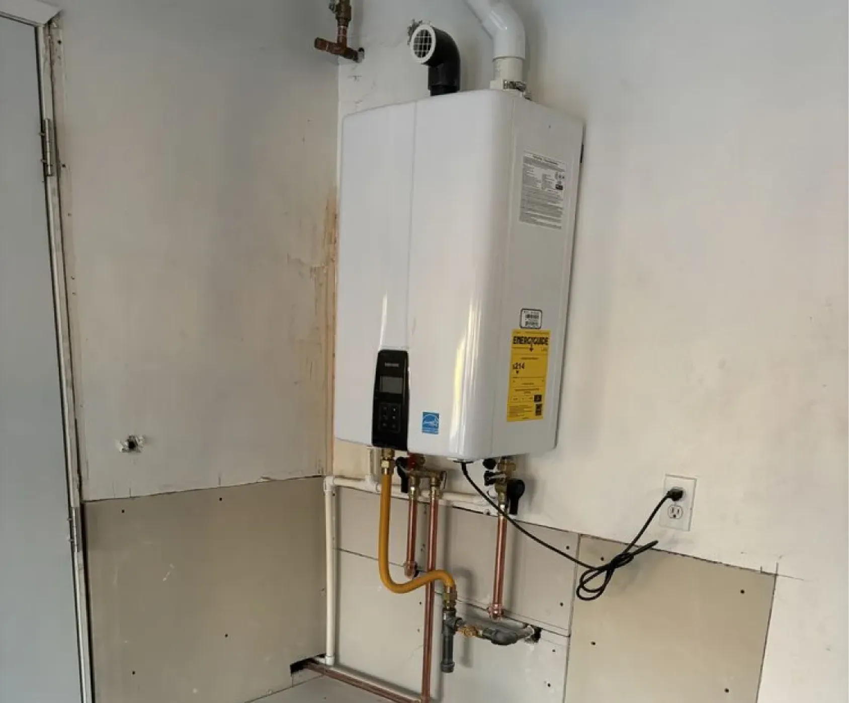 A tankless water heater mounted on a wall with pipes and a power cord connected.