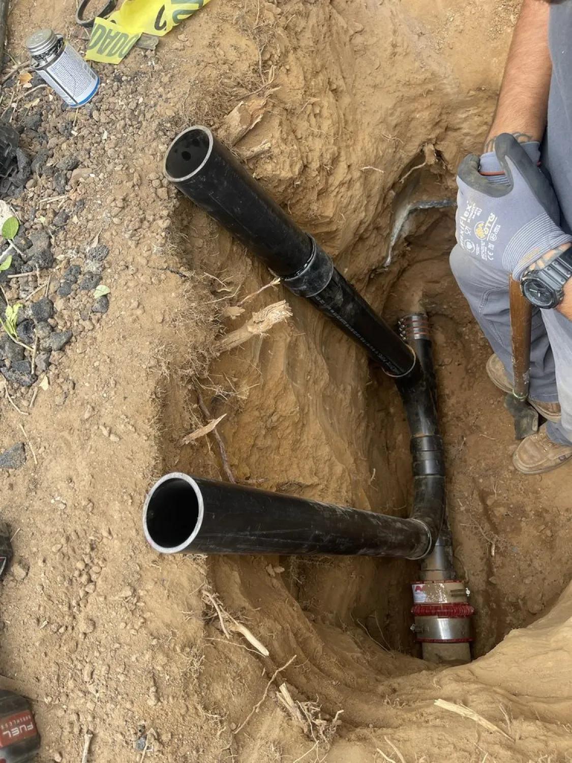 why-choose-1 Two black pipes in a dirt trench, with a person holding a tool and wearing gloves on the right side.